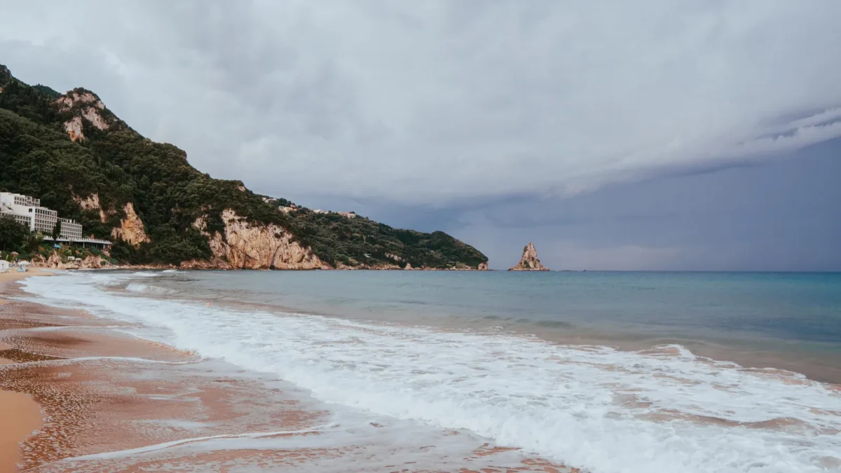 Zandstrand op Corfu met golven, groene kliffen en een bewolkte lucht